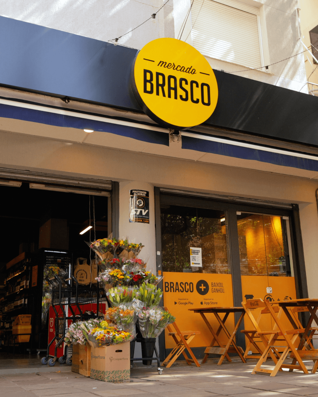 Fachada do Brasco Florêncio: toldo azul-marinho com a placa amarela do Mercado Brasco, buquês de flores na entrada e mesas de madeira na calçada.