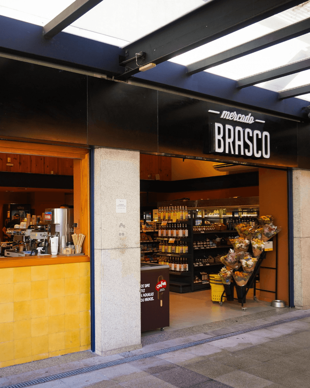 Entrada do Brasco no Viva Open Mall: letreiro Mercado Brasco, balcão de café à esquerda, prateleiras da mercearia e carrinho de flores.